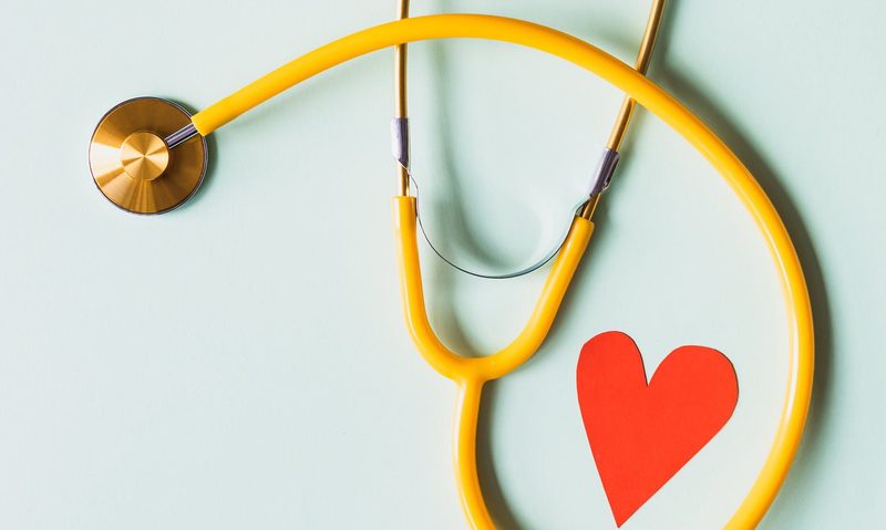 Stethoscope wrapped around a small paper red heart on a light blue background