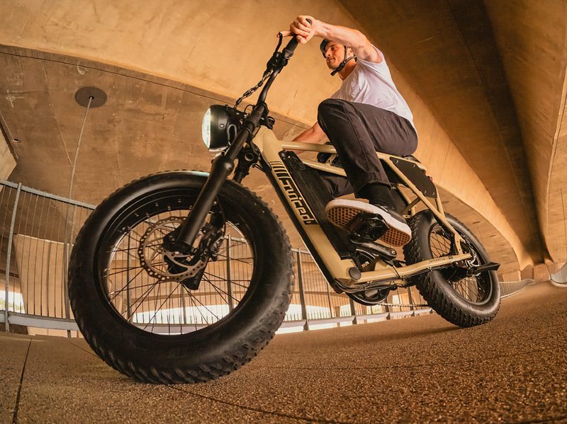 Front left three-quarter view looking up at a rider with helmet on a Juiced Bikes Scrambler X2 riding in a covered structure that looks like a parking garage.