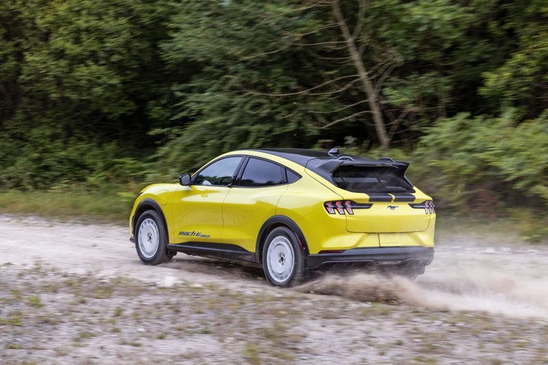 Mustang Mach-E Rally left rear three-quarter view driving away on a dirt road in the woods.