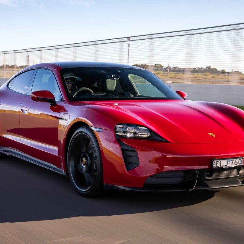 Red Porsche Taycan Turbo S EV right front three-quarter view driving on a highway with a small body of water in the background.