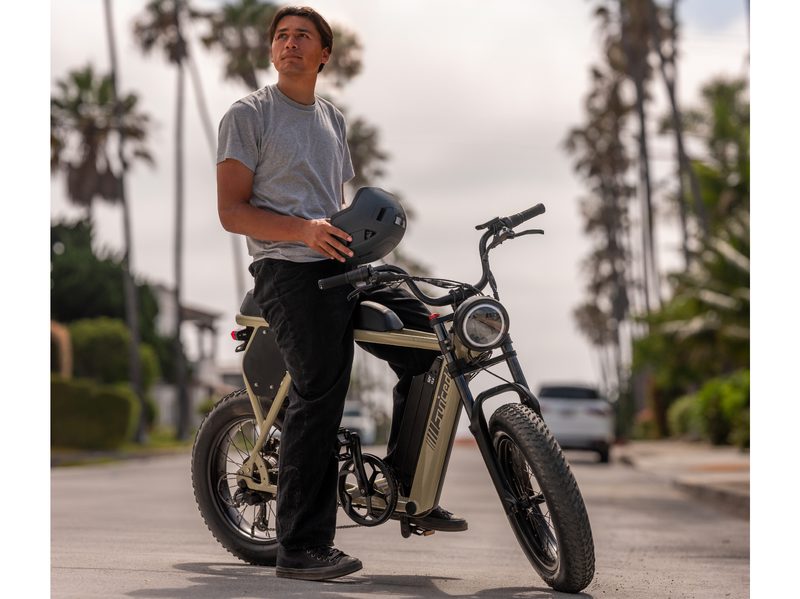 Right front three-quarter view of a Juiced Bikes Scrambler X2 with a rider holding a helmet sitting on the bike in a side street with palm trees and ocean in the background.