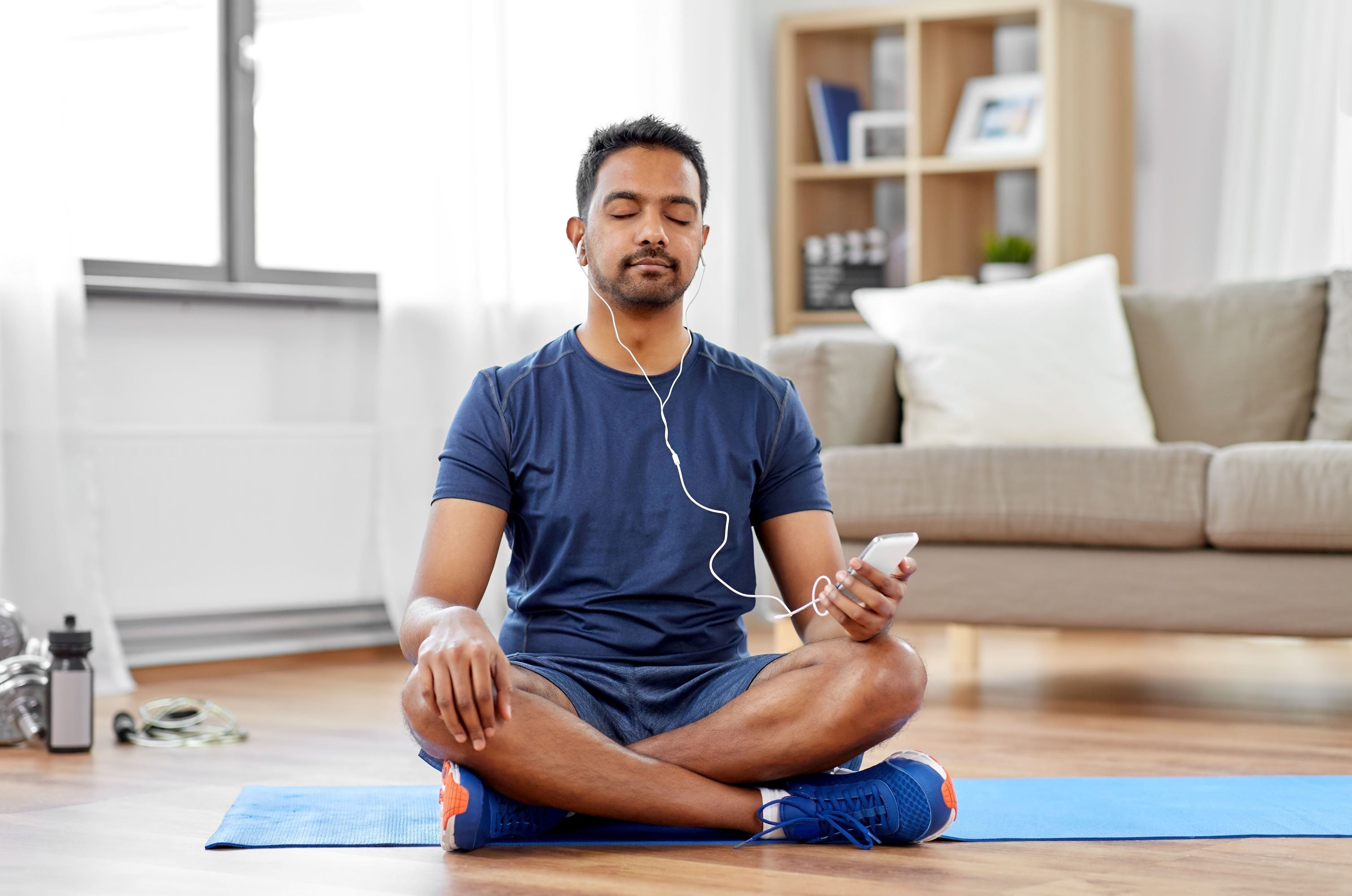 man wearing blue shirt sitting on yoga mat on wooden floor at home with smartphone listening to music with headphones yoga app