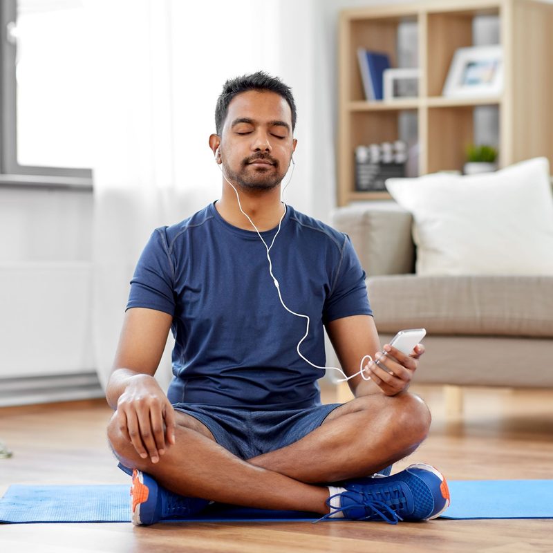 Man wearing blue shirt sitting on yoga mat with phone listening to music meditation
