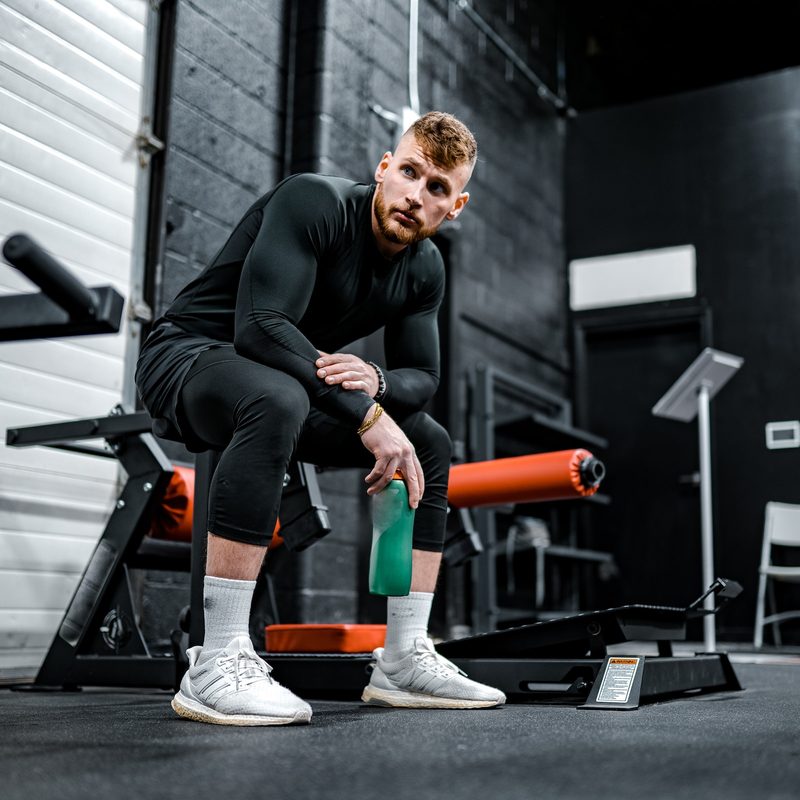 Guy sitting on bench at gym holding water flask wearing black with white sneakers