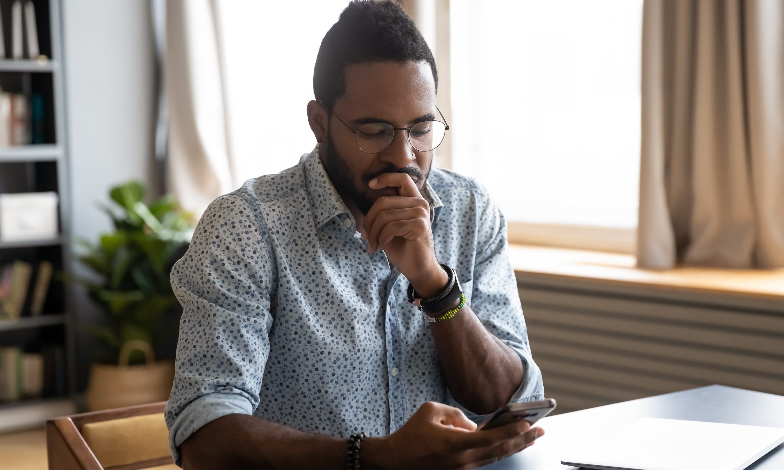 Confused African guy holding smart phone feels concerned thinking over received message.