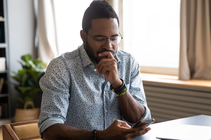 Confused African guy holding smart phone feels concerned thinking over received message.