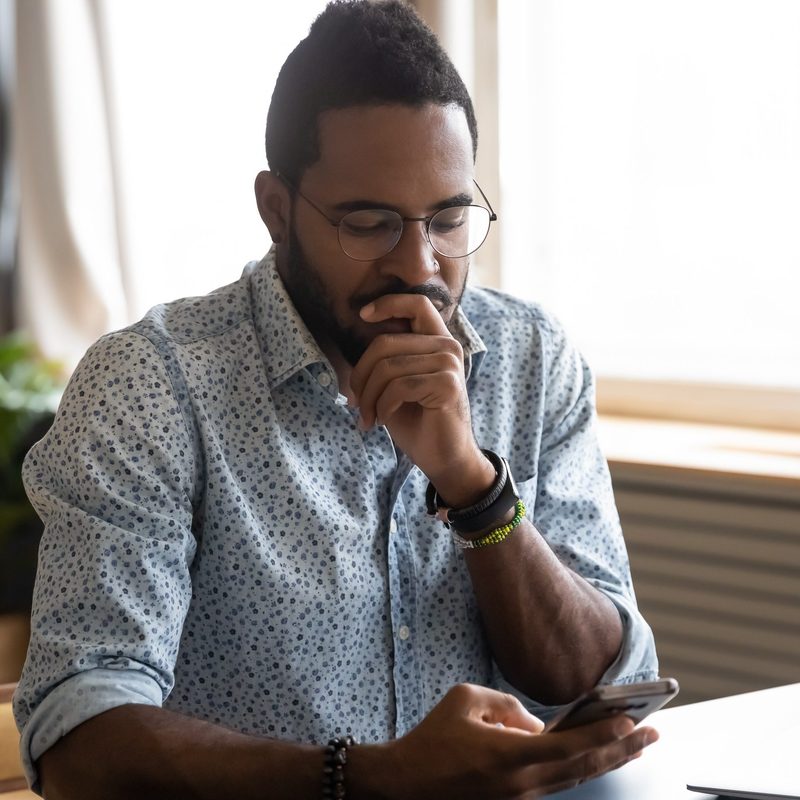 Confused African guy holding smart phone feels concerned thinking over received message.