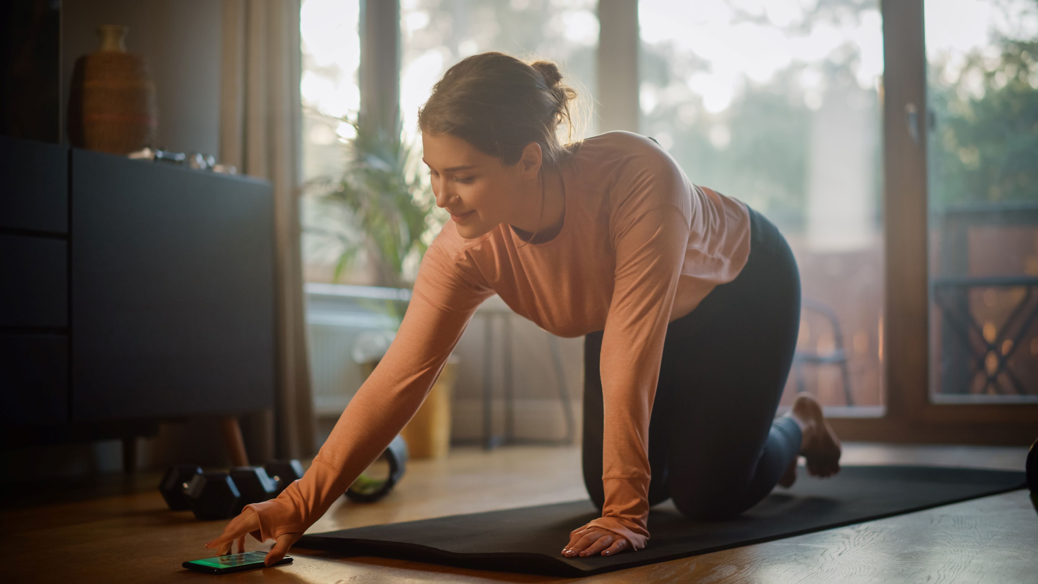 girl on yoga mat using smartphone wearing pink shirt and black leggings in front of window
