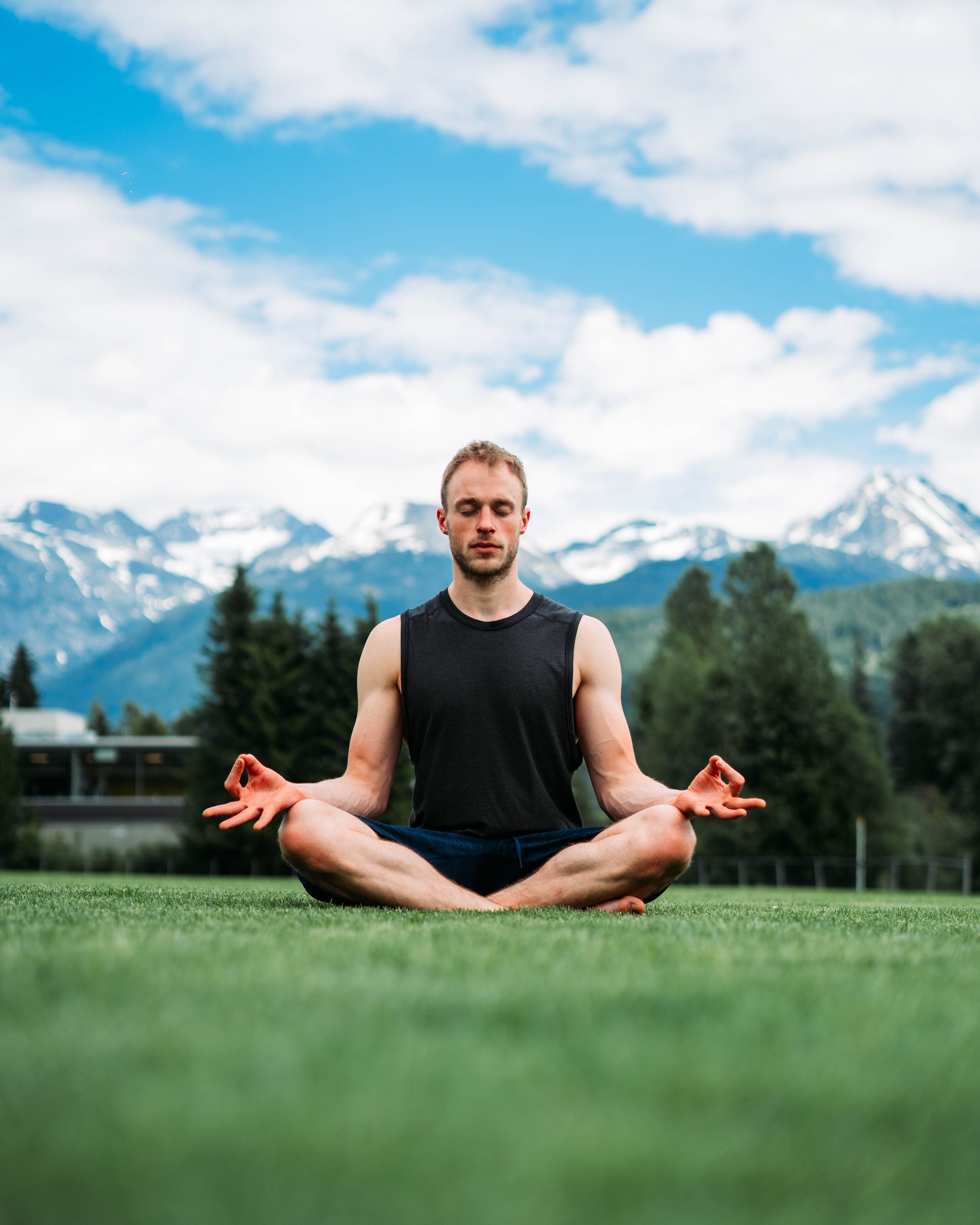 man sitting outside on the grass under a blue sky in a cross legged yoga pose