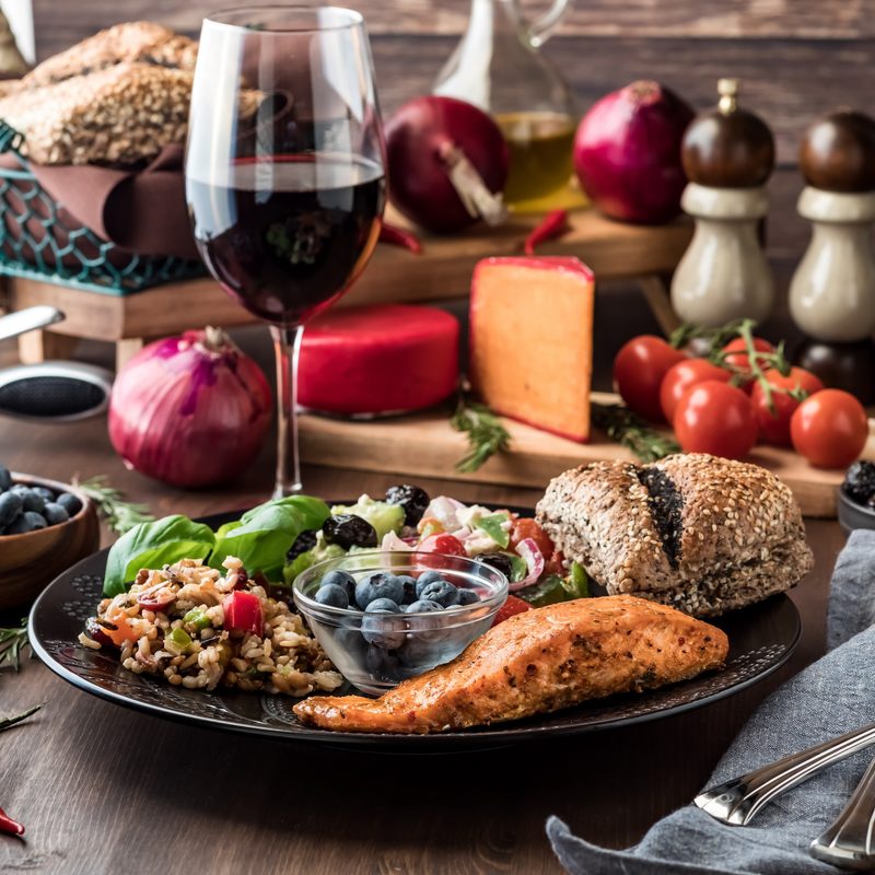Mediterranean meal of greek salad, seven grain salad, salmon fillet and a small bowl of blueberries, surrounded by Mediterranean diet foods.