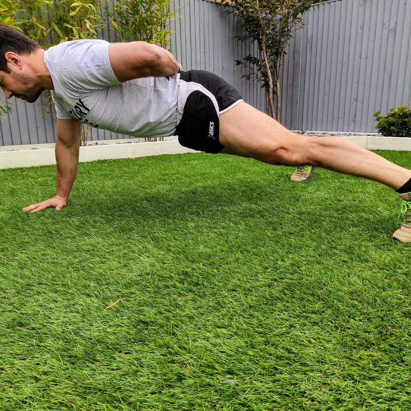 Man in plank pose one hand behind back on grass outside wearing shorts and shirt