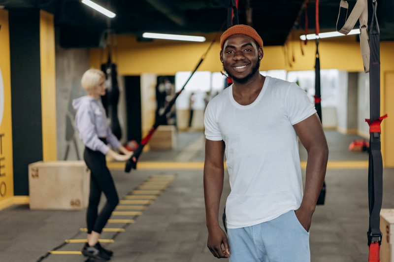 Man smiling to the camera with woman working out behind him
