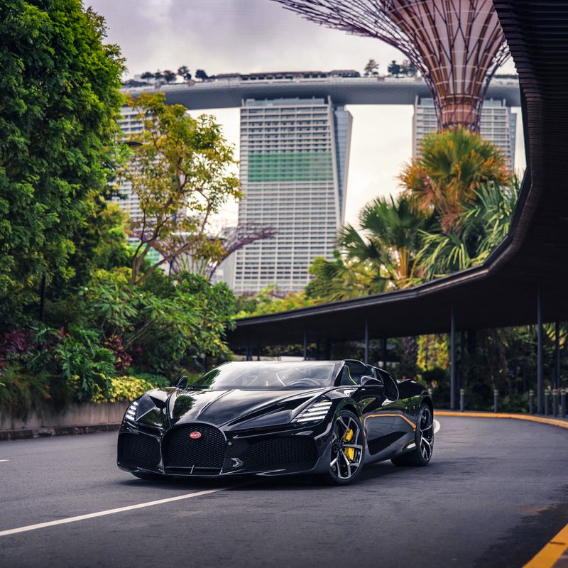 A black Bugatti W16 Mistral Roadster driving on a street in Singapore with heavy vegetation beside the road and modernistic structures overhead.