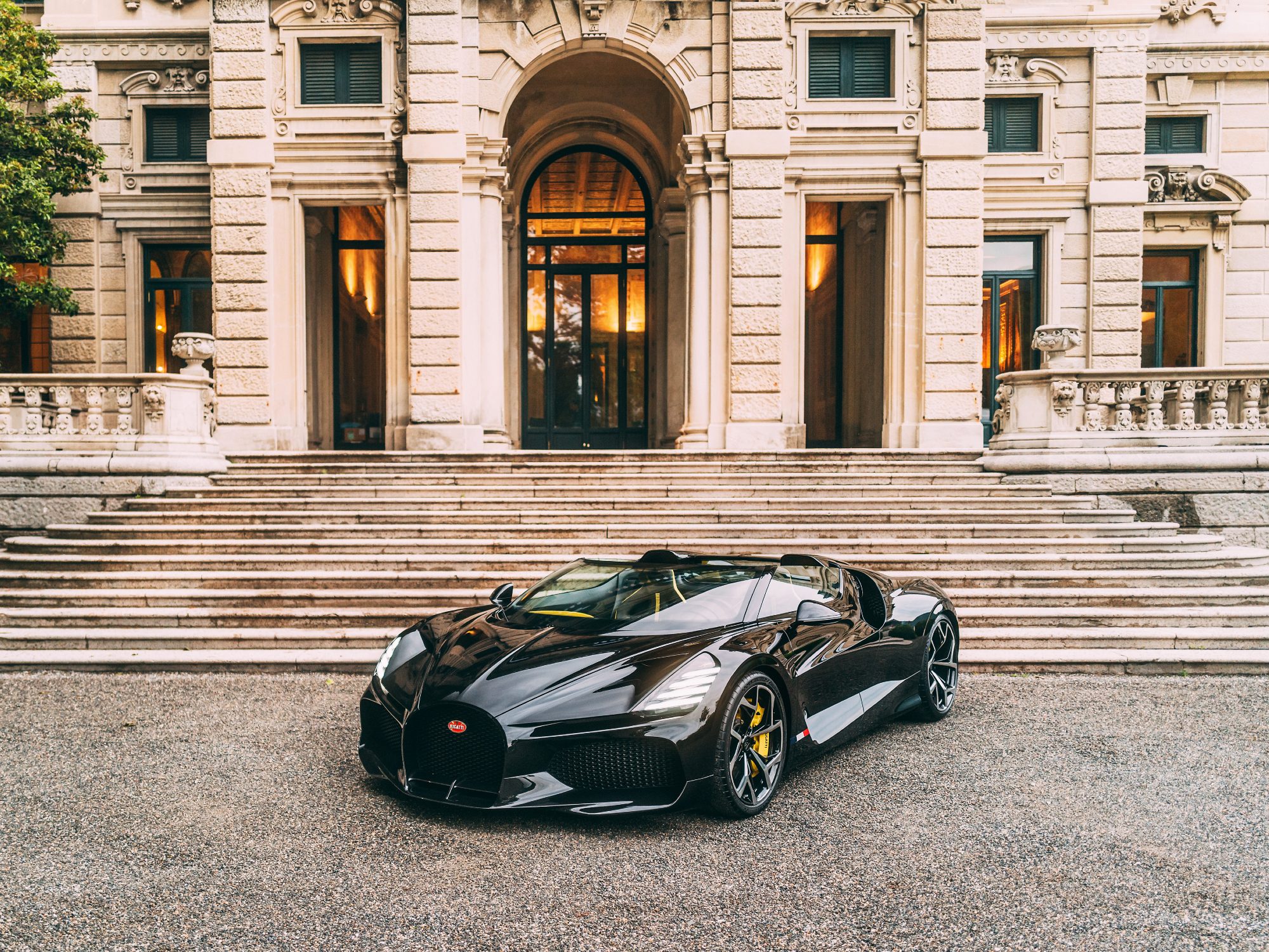 A black Bugatti W16 Mistral Roadster parked facing out at the bottom of a wide light stone entry stairway to an elaborate light stone building.
