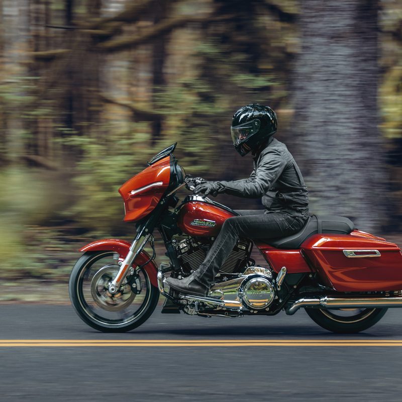 A person with a gray riding suit and black helmet riding a 2024 Harley-Davidson Street Glide on a two-lane street with a forest in the background.