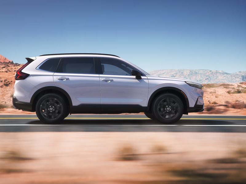 A white 2024 Honda CR-V Hybrid right side shot driving on a paved road with desert landscape in the background.