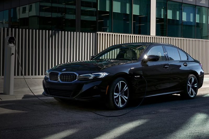 Black 2024 BMW 330e PHEV plugged into a charger parked on the side of a city street.