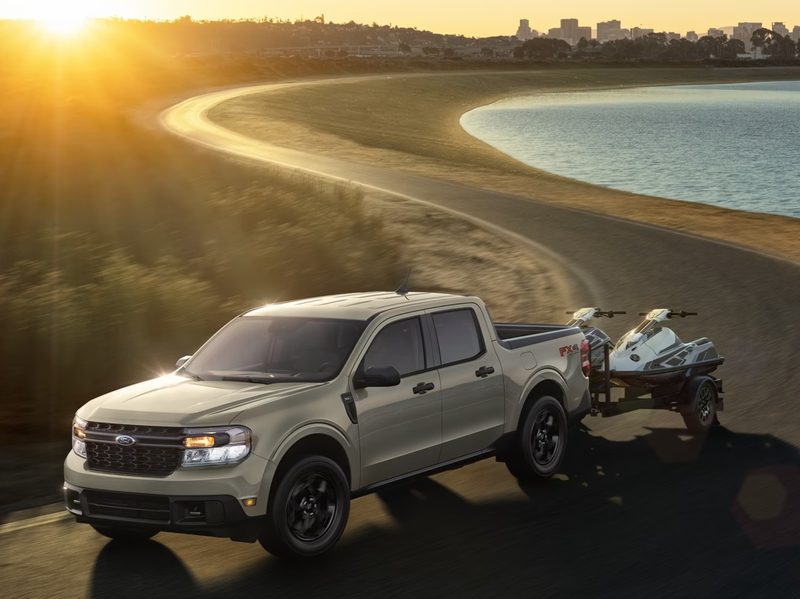 Light tan 2024 Ford Maverick hybrid pickup truck towing two jet skis on a small trailer on a curved road near a lake.