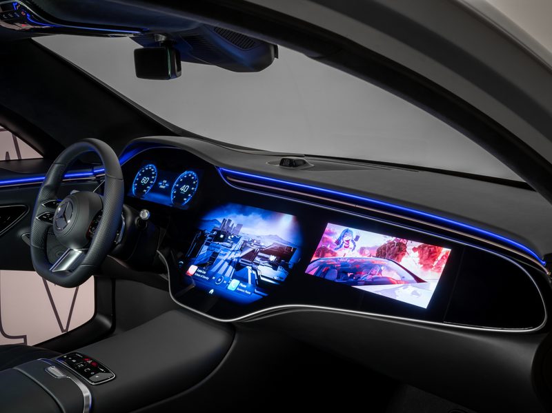 Mercedes-Benz MBUX photo from passenger seat showing steering wheel with driver information screens, large center infotainment display, and 3D graphics on passenger screen.