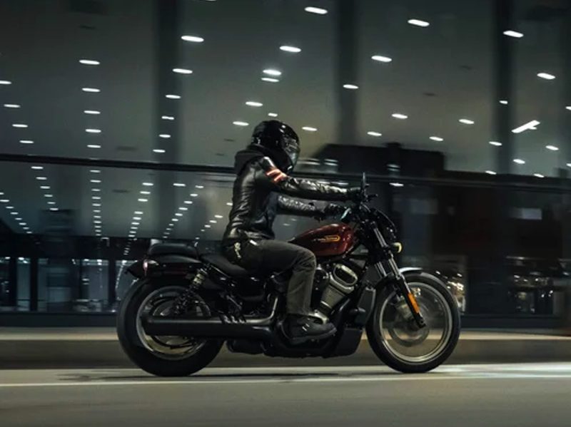 Rider on a 2024 Harley-Davidson Nightster cruiser motorcycle driving by a building with all-glass walls.