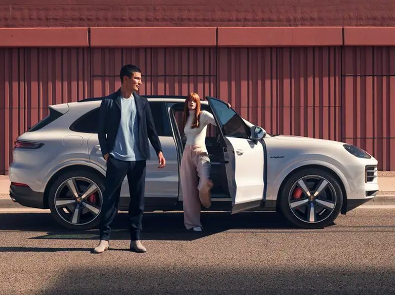 Two people and a 2024 Porsche Cayenne E-Hybrid parked on a city street with high brick-red fencing.