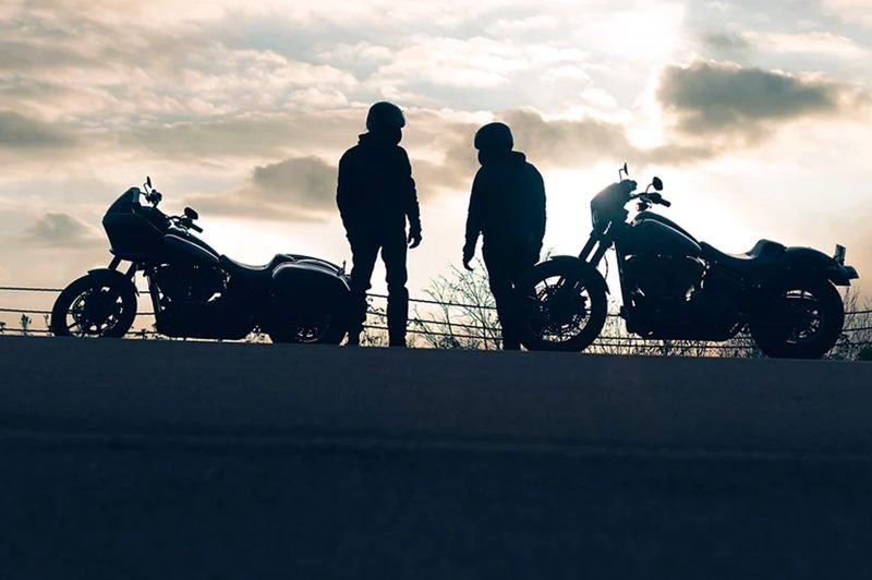 Two suited-up riders wearing helmets standing with 2024 Harley-Davidson Low Rider ST motorcycles with the setting sun in the background.