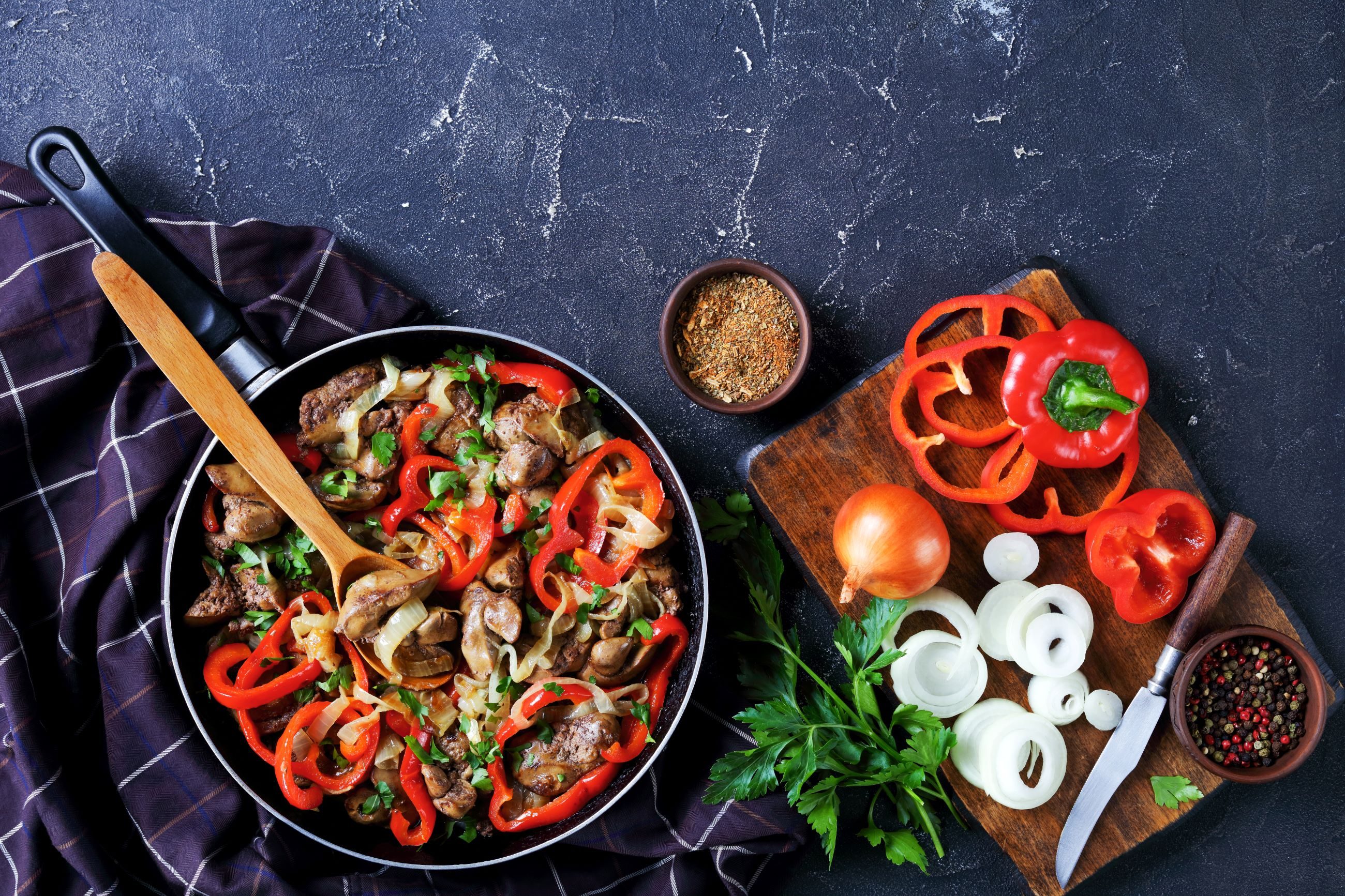 liver with peppers onions in skillet next to cutting board with onions and herbs on table
