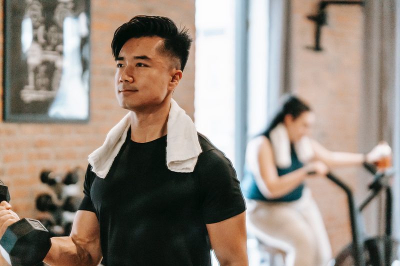 Man at the gym with a towel around his neck and woman riding stationary bike behind him.
