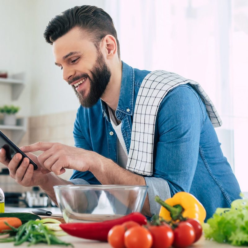 Happy bearded man is using his smartphone while he preparing a meal