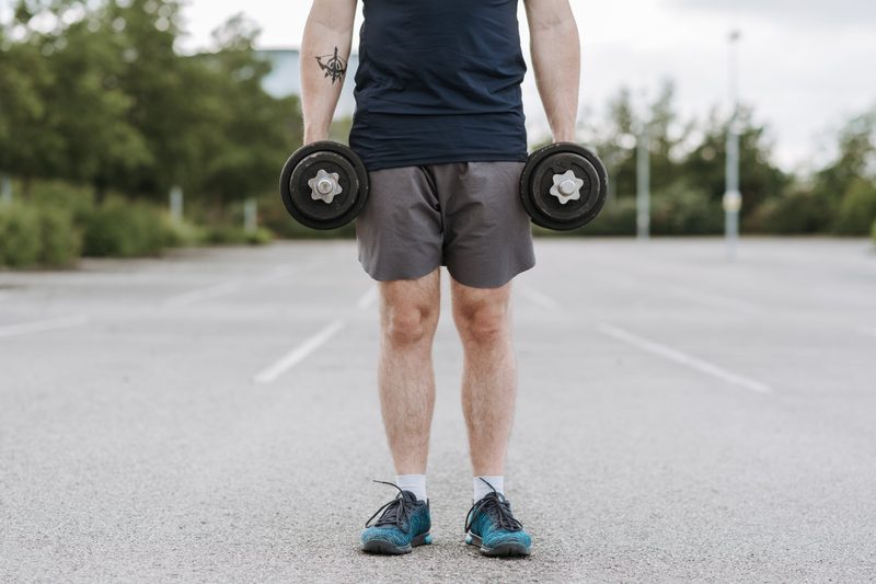 Person standing on asphalt holding a dumbbell in each hand.