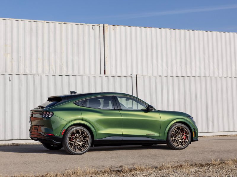 2024 Ford Mustang Mach-E Bronze shot taken of right side with the car parked in front of two gray stacked shipping containers.