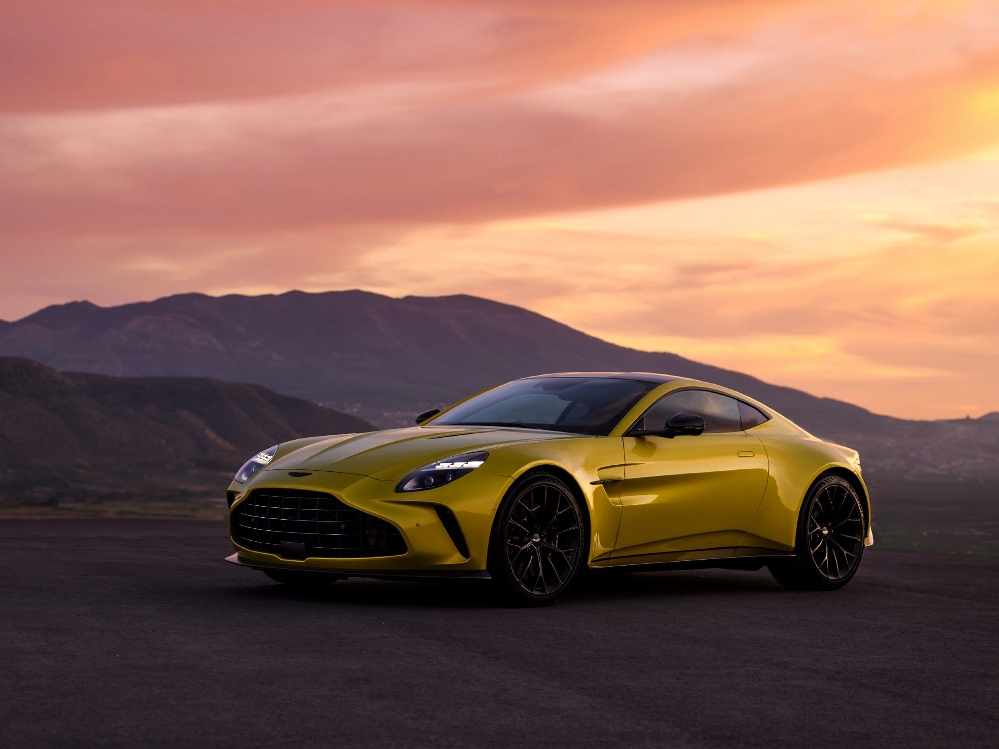 Aston Martin Vantage sports car left front three-quarter view parked on black surface with black hills and sunset skies.