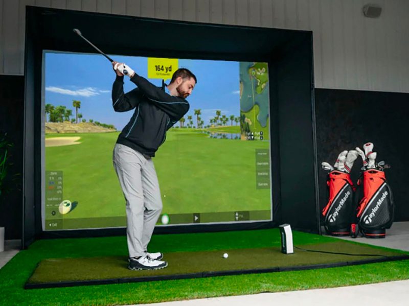 A man swings a golf club on the OptiShot Golf In a Box 4 golf simulator.