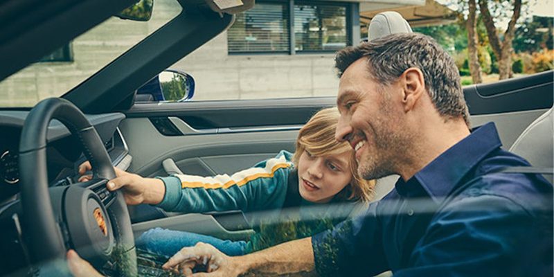 A man and boy in a Porsche with the man smiling as the boy touches the steering wheel.