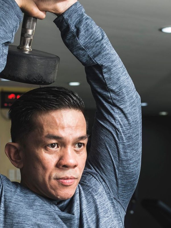 man in gym wearing long sleeve blue shirt doing overhead tricep extension exercise with weight