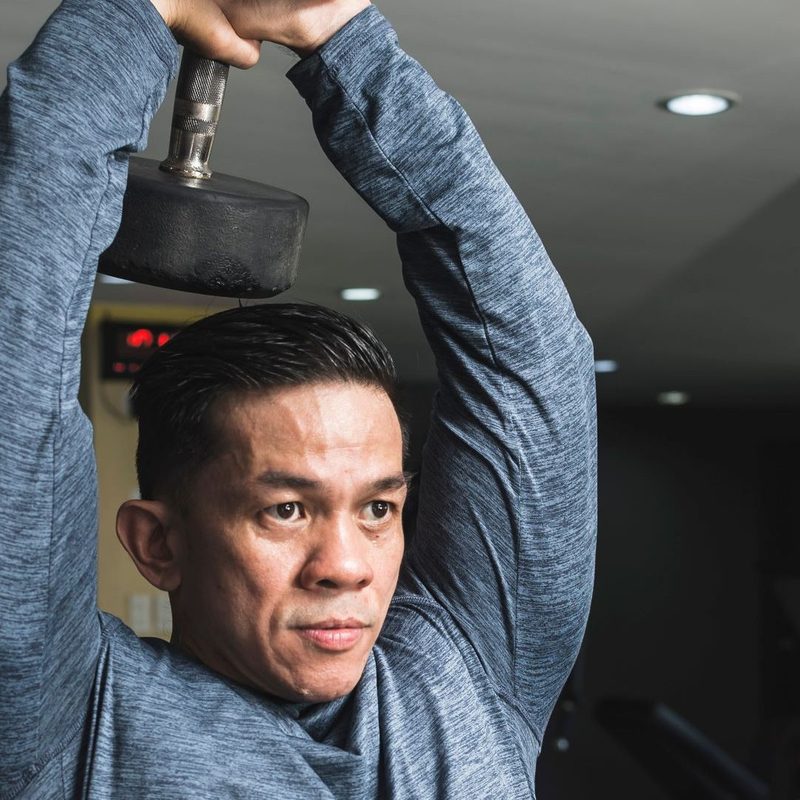 Man in gym wearing long sleeve blue shirt doing overhead tricep extension exercise with weight