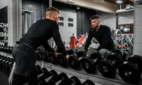 Man in gym wearing black looking in mirror lifting up weights next to weight rack