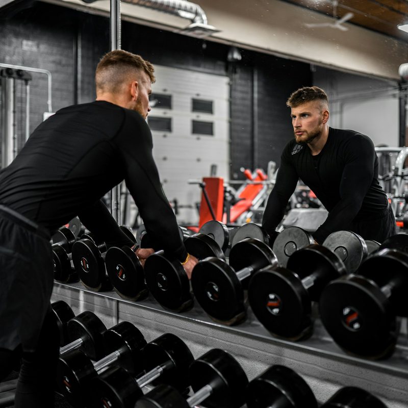 Man in gym wearing black looking in mirror lifting up weights next to weight rack