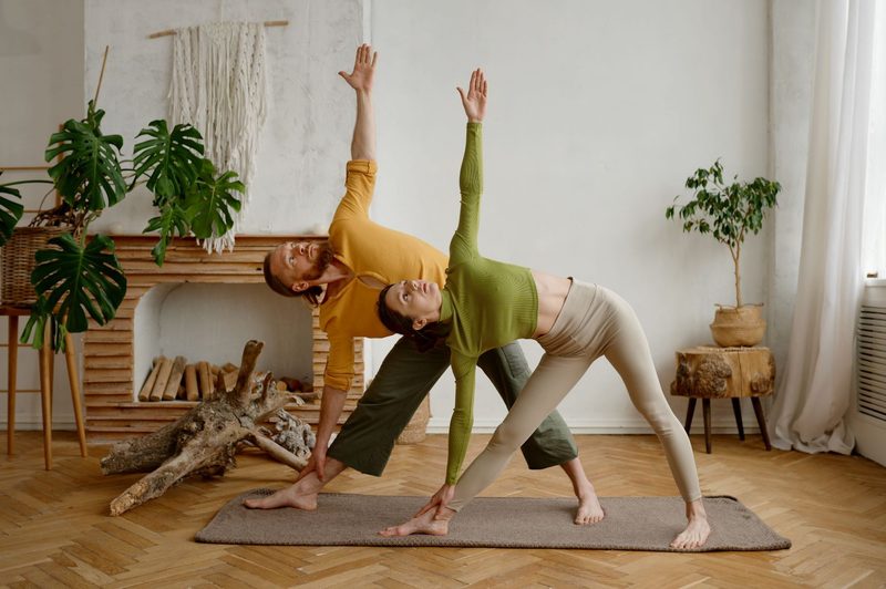 Man and woman doing triangle yoga pose on mat indoors on wooden floor