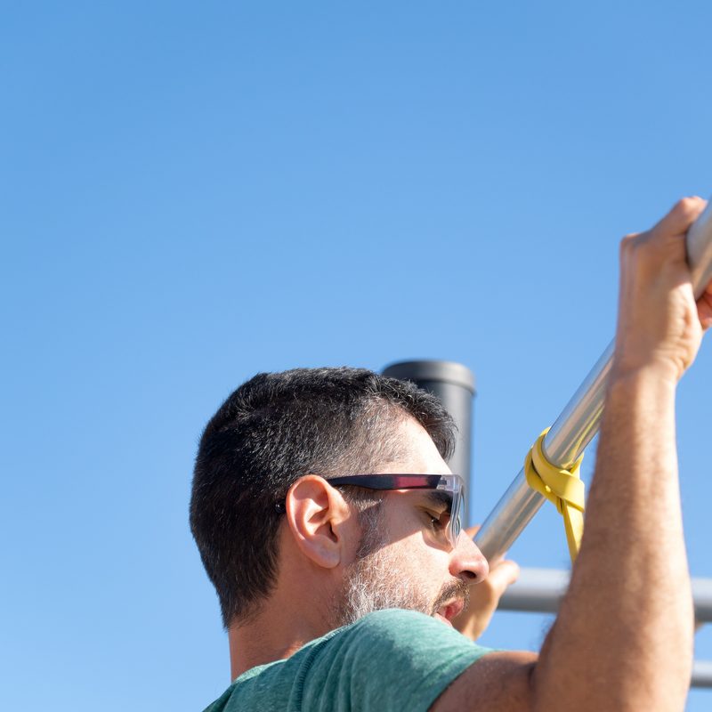 bearded man with sunglasses seen from behind training his back by doing pull-ups on a barbell with an elastic fitness band in an outdoor gym.