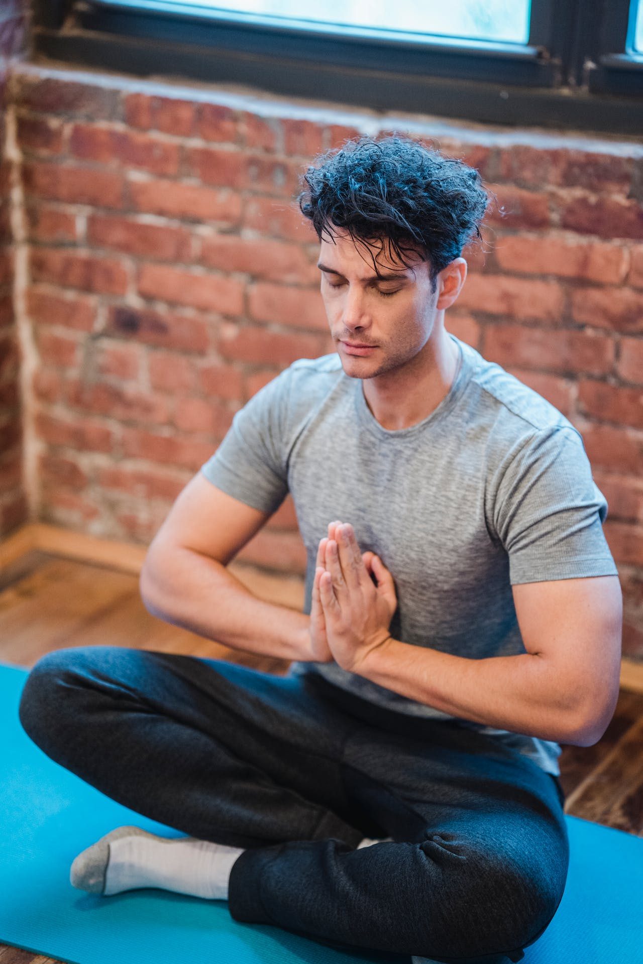 man seated indoors by brick wall on a yoga mat doing seated yoga pose