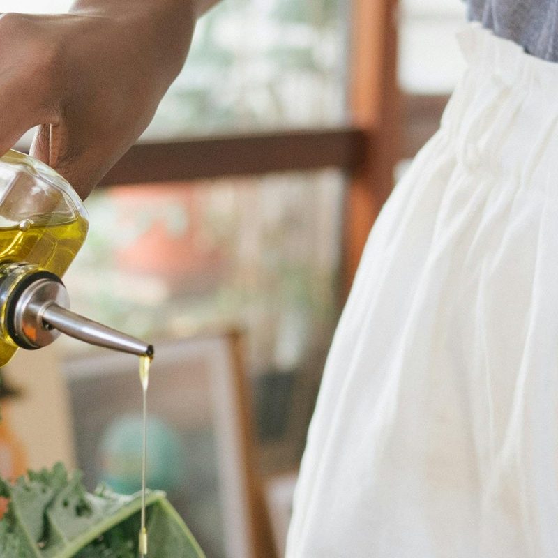 Cooking pouring olive oil onto black pan with veggies and herbs on wooden table