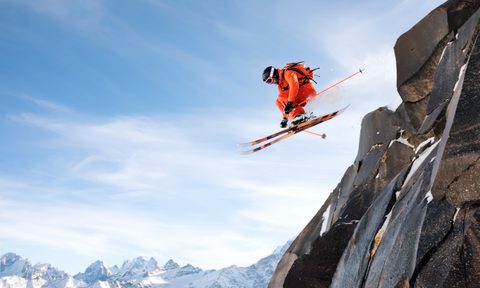 A professional skier makes a jump-drop from a high cliff against the blue sky leaving a trail of snow powder in the mountains. Caucasian ridge in the background. Photo from the slopes of Mount Elbrus. The concept of extreme sports and recreation in the mountains in winter. Copy the space