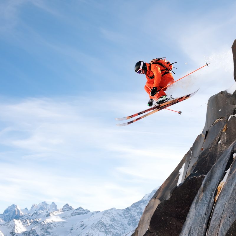 A professional skier makes a jump-drop from a high cliff against the blue sky leaving a trail of snow powder in the mountains. Caucasian ridge in the background. Photo from the slopes of Mount Elbrus. The concept of extreme sports and recreation in the mountains in winter. Copy the space
