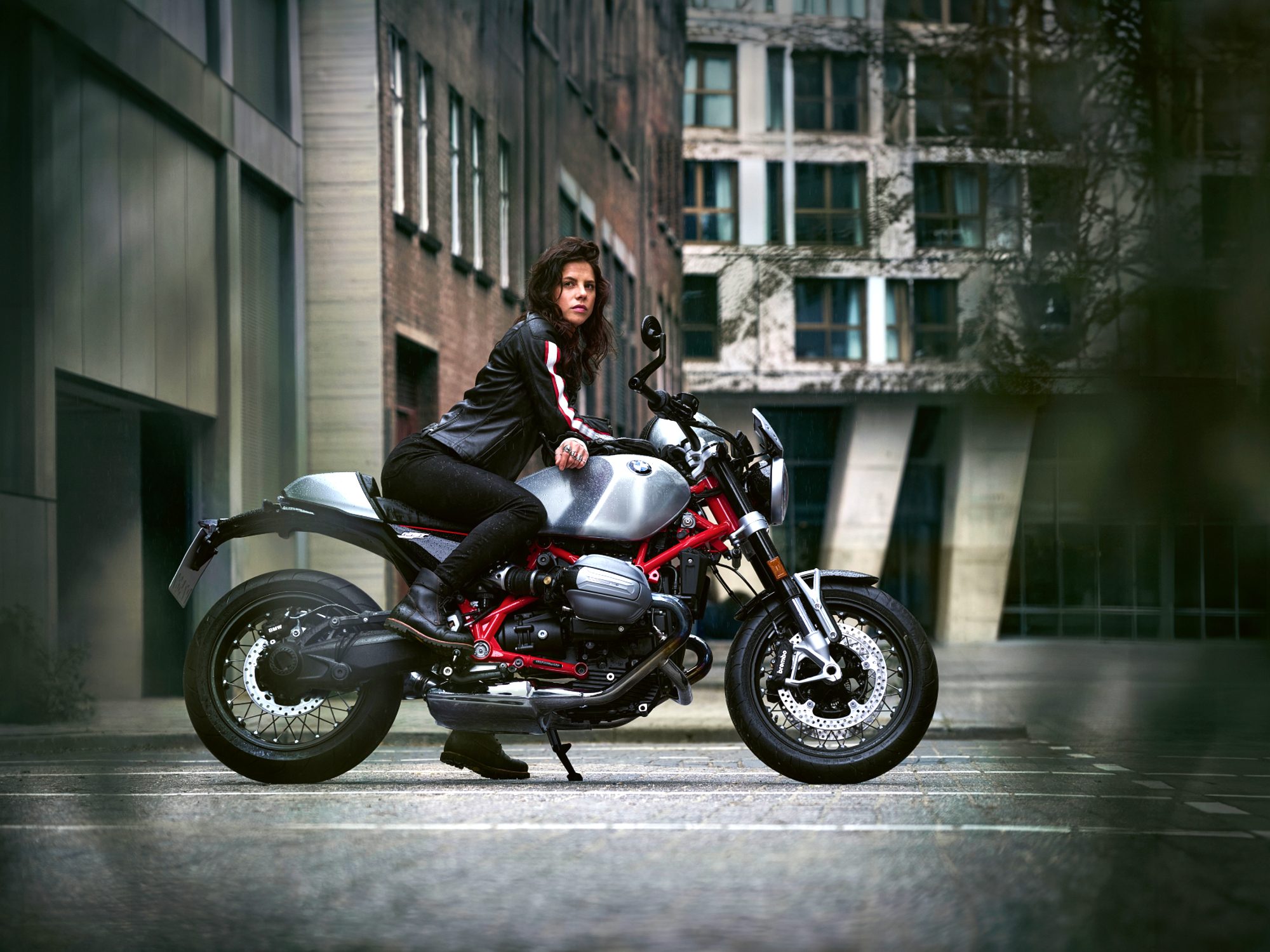 A rider sitting on a parked 2024 BMW R 12 NineT motorcycle with helmet off and arms crossed over tank with office buildings in the background.