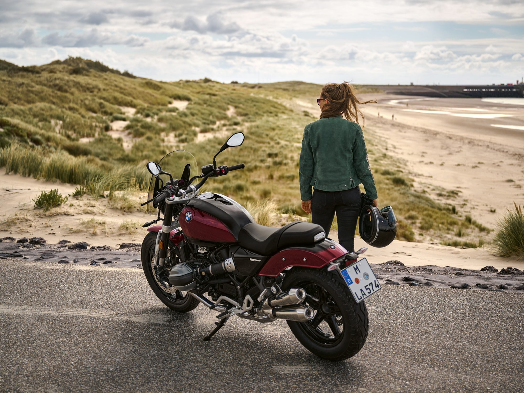 A rider standing on the far side of a oarked 2024 BMW R 12 NineT motorcycle looking a beach and dunes.