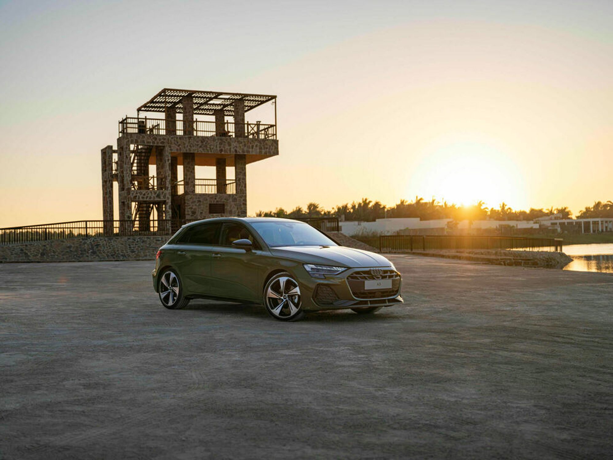 Green Audi A3 Sportback parked on a beach at sundown with a lifeguard stand and water in the background.