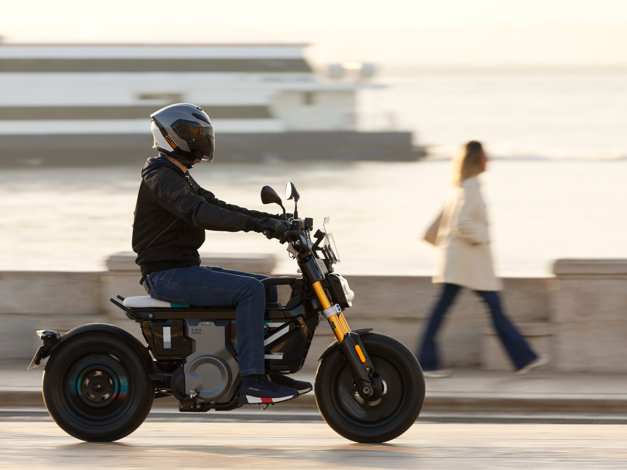 Helmeted rider traveling on a city street on a 2024 BMW CE 02 with a two-deck sea ferry docked on a waterway in the background.