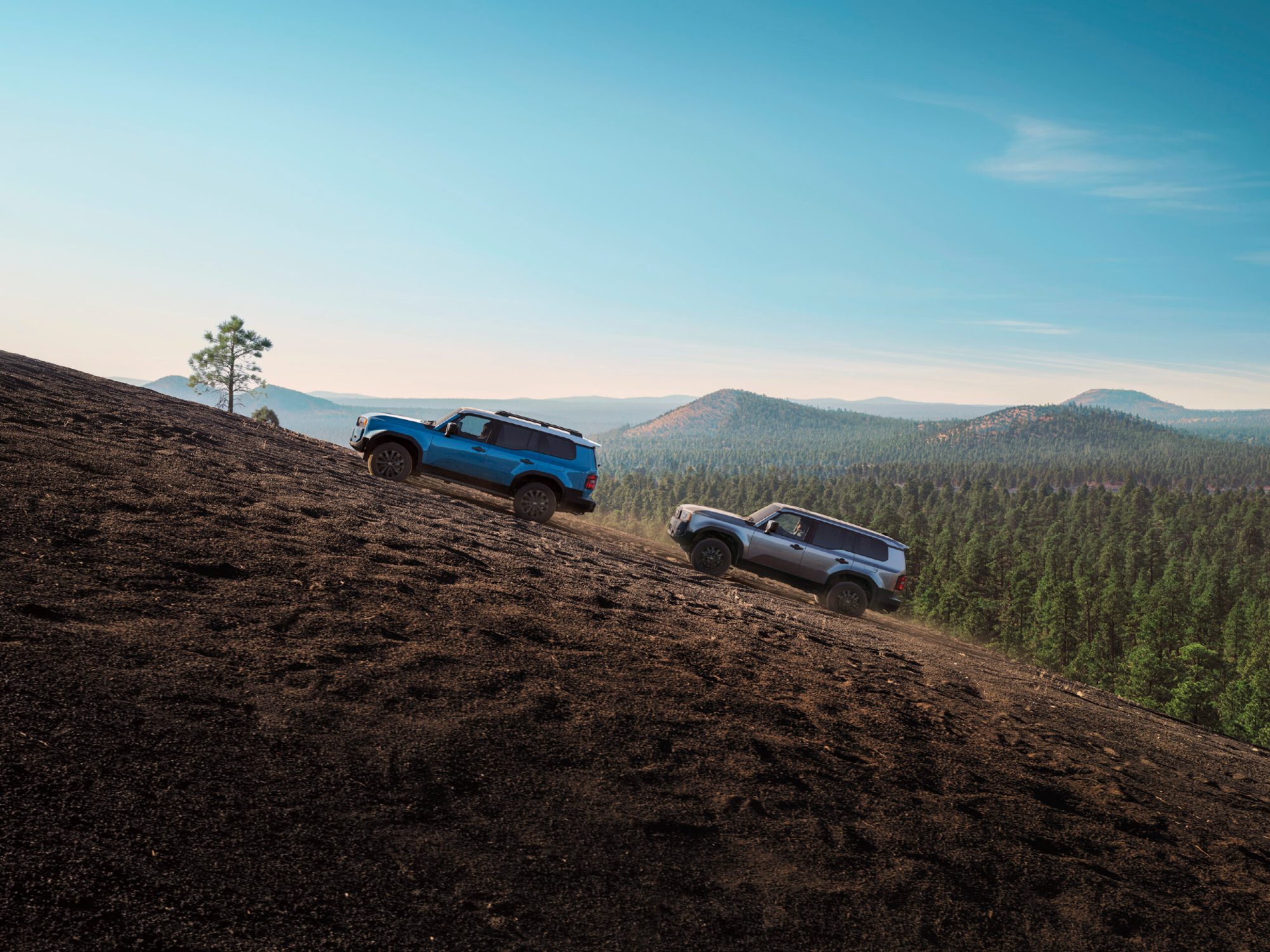 Two 2024 Toyota Land Cruisers driving up a steep slope with mountains in the distance.