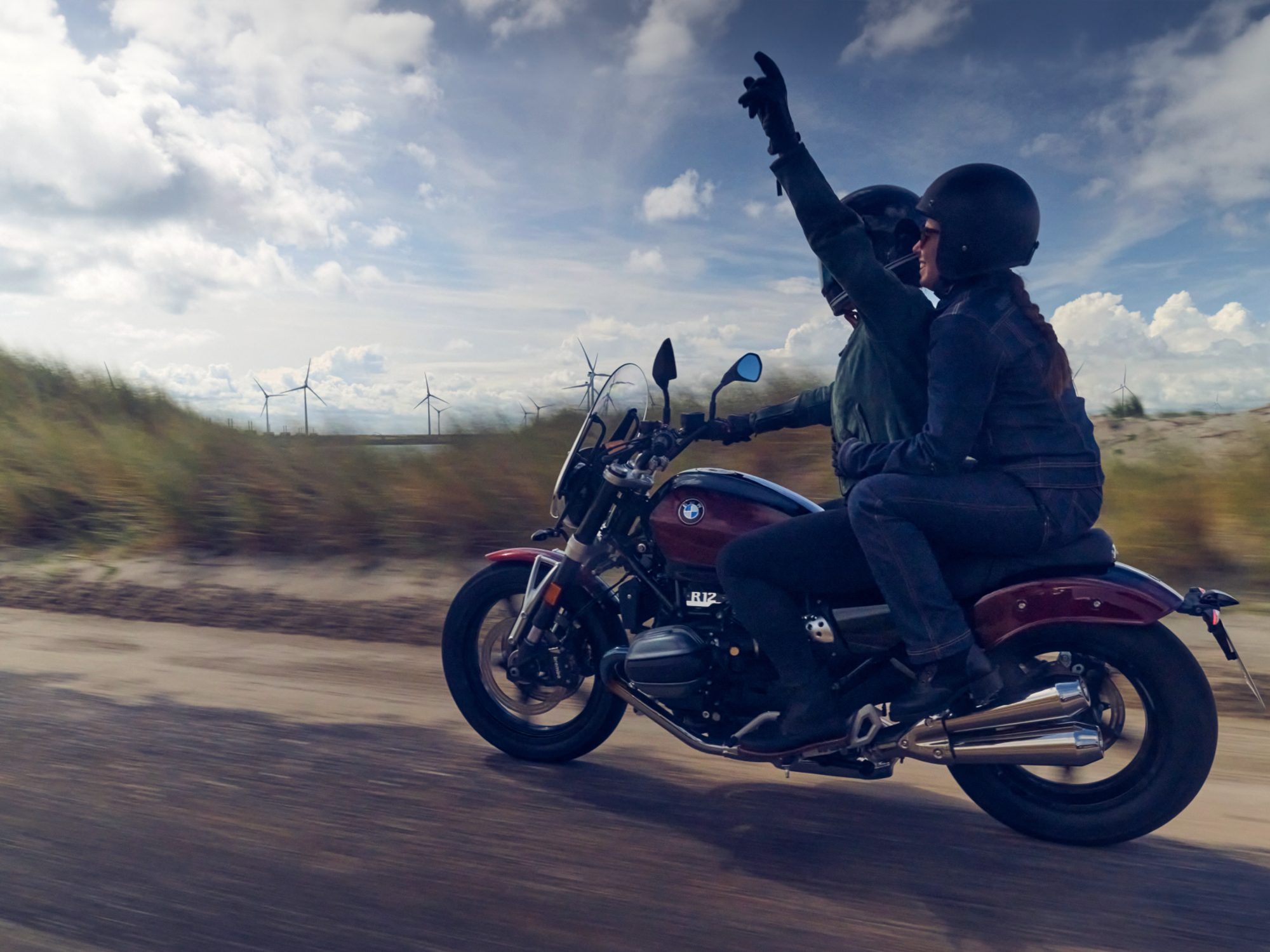 Two riders on 2024 BMW R 12 on a dirt road with grassy fiedls and dune in the back ground.